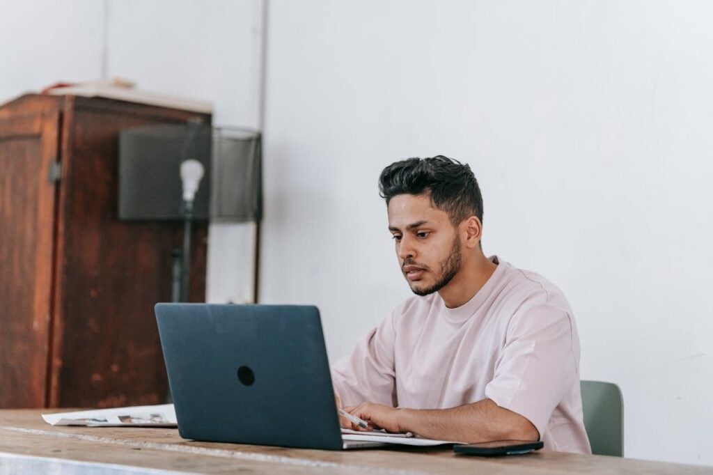 Um homem de barba e cabelos escuros, vestindo uma camiseta rosa claro, sentado em uma escrivaninha. Ele trabalha em um laptop azul escuro e segura uma caneta, parecendo anotar informações enquanto olha atentamente para a tela.