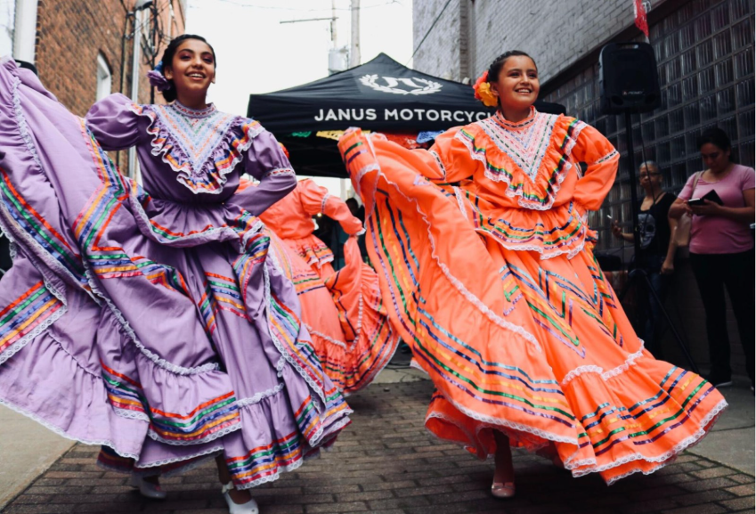 Foto de duas mulheres com vestidos longos, um roxo e outro laranja, e dançando uma dança típica de sua cultura 