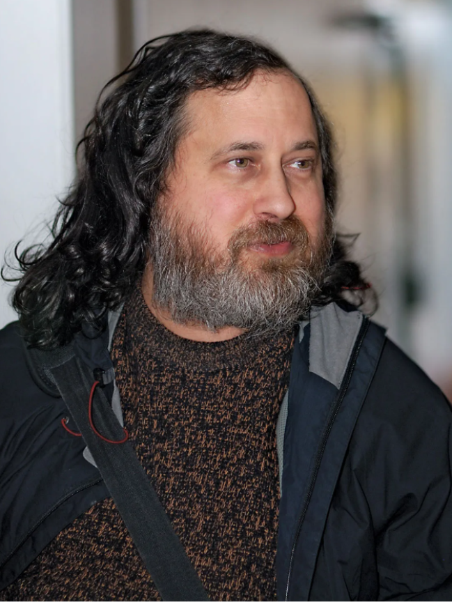 Homem de cabelo longo e barba grisalha olhando para o lado, usando casaco escuro e suéter marrom. 