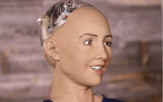 Vídeo em loop que mostra a robô Sophia, que funciona com inteligência artificial. A robô Sophia apresenta uma pele emborrachada, a cor da pele é clara e não tem cabelos. A parte de trás da cabeça deixa à mostra os circuitos elétricos do robô. A Sophia está sorrindo e com os olhos arregalados, ela está com a cabeça virada para frente e vira lentamente à direita, olhando diretamente para a câmera como ar assustador e engraçado ao mesmo tempo. 
