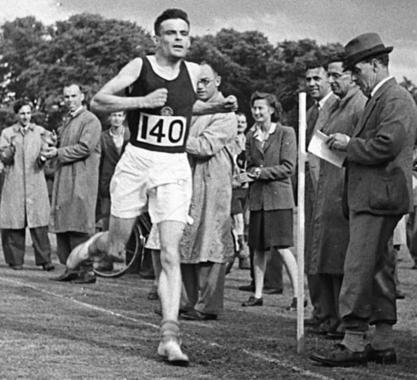 Imagem de Alan Turing, um homem caucasiano e de cabelos escuros, com trajes de corredor e o número 140 na camiseta preta, correndo e várias pessoas ao redor observando sua chegada. 