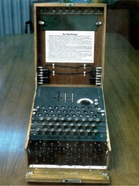 Imagem que mostra a máquina Enigma. Uma máquina eletromecânica envolvida por um caixote de madeira similar a uma mala. A máquina está aberta, seu interior é preto e apresenta um teclado com letras, acima do teclado há alguns sinalizadores das letras que acendiam após as teclas serem selecionadas. Abaixo do teclado há os rotores à mostra. Na tampa aberta há uma mensagem colada na caixa. 