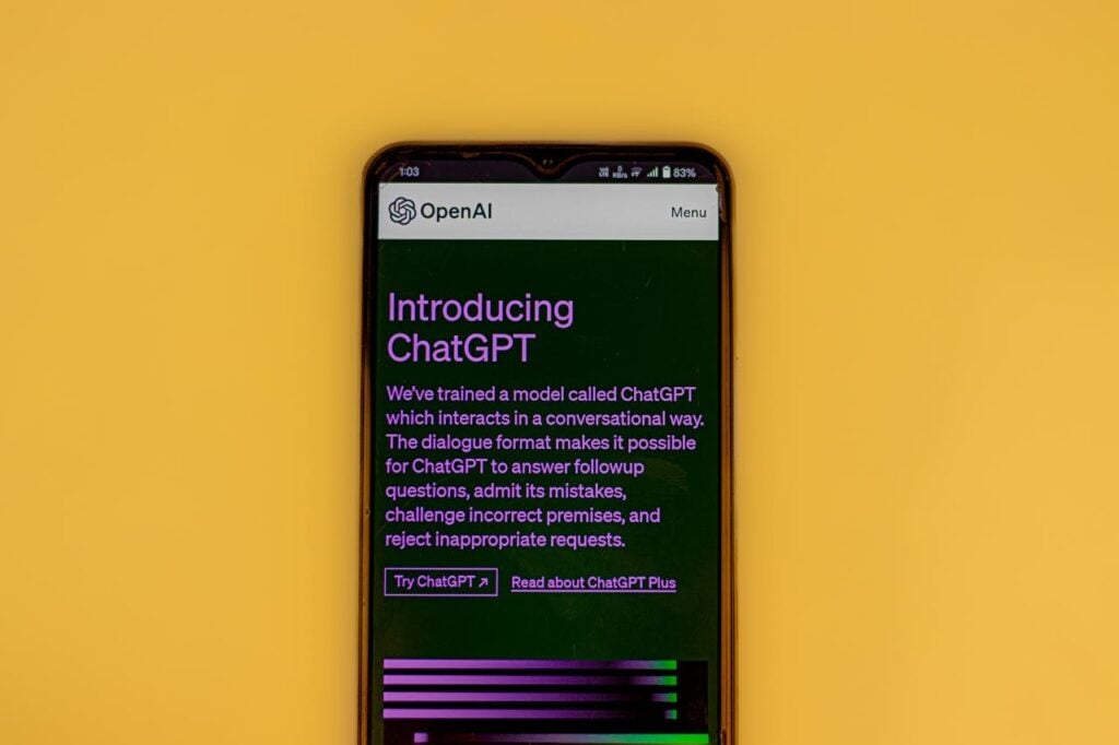 Um smartphone centralizado sobre um fundo amarelo vibrante. A tela do aparelho exibe o site oficial da OpenAI com o título "Introducing ChatGPT" sobre um fundo verde escuro, detalhando que o modelo foi treinado para interagir de forma conversacional.