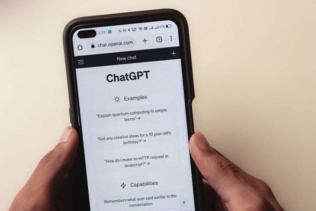 Visão próxima de mãos segurando um smartphone que exibe a interface de um "New chat" (nova conversa) do ChatGPT. São visíveis os blocos de "Examples" (Exemplos), como pedidos para explicar computação quântica, e a seção de "Capabilities" (Capacidades).