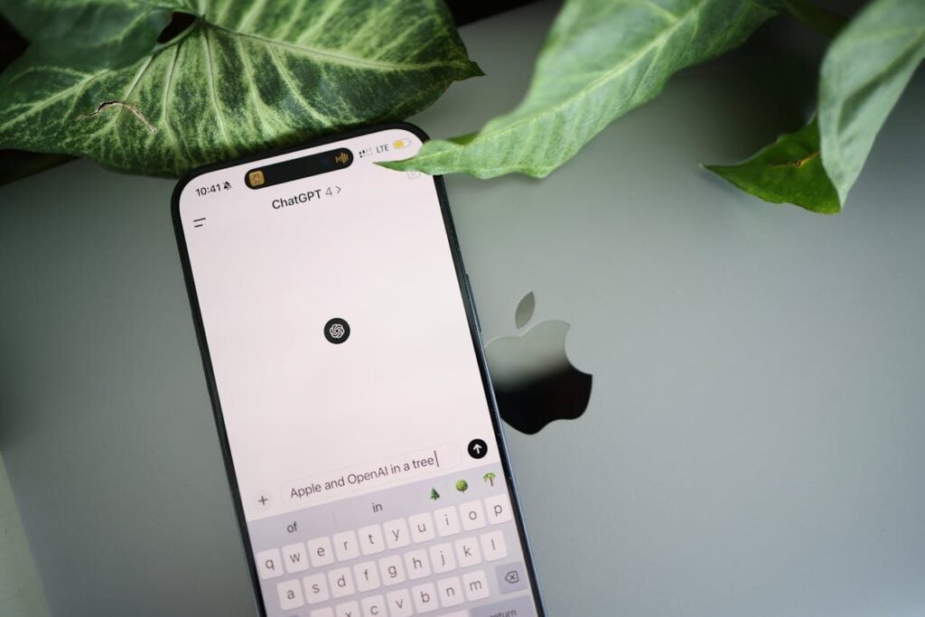 Um smartphone repousado sobre a superfície prateada de um laptop (com o logotipo da Apple parcialmente visível) e ao lado de folhas verdes de uma planta. A tela do celular mostra o campo de digitação do ChatGPT com o prompt "Apple and OpenAI in a tree" sendo escrito.