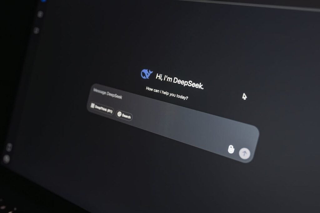 Fotografia em close de um monitor de computador em ambiente escuro, exibindo a interface da inteligência artificial DeepSeek. Na tela preta, destaca-se o texto de saudação "Hi, I'm DeepSeek. How can I help you today?" logo acima de uma barra de pesquisa.