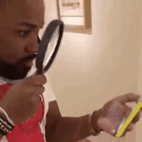 Homem com expressão séria segurando uma lupa próxima ao rosto enquanto olha para a tela do celular com atenção. Ele move a lupa para tentar enxergar melhor, sugerindo que a tela tem algo pequeno ou difícil de ler.