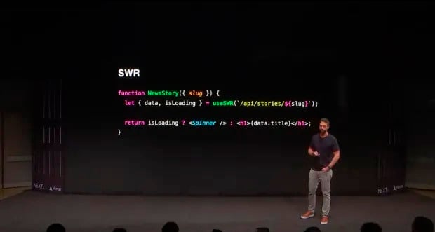 Sam Selikof está de pé em um palco ao lado de uma projeção de uma função de código de programação chamada 'NewsStory'. O código mostra o uso do gancho 'useSWR' para buscar dados de uma API com uma variável 'slug'. Ele verifica se o conteúdo está carregando e, caso esteja, mostra um componente 'Spinner'; se não, exibe o título da história em um elemento de cabeçalho 'h1'. O fundo é escuro e o logo da NEXT é visível no canto inferior esquerdo.