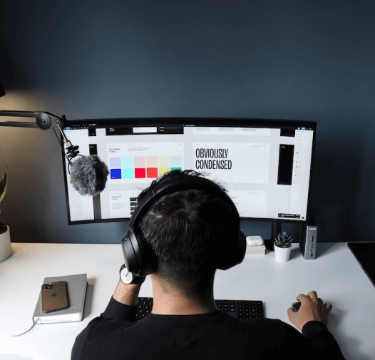Imagem mostra uma pessoa de costas, usando fones de ouvido e trabalhando em um monitor que exibe um design com cores e texto. A pessoa está sentada em uma mesa branca que contém um teclado, mouse e celular. O ambiente tem uma parede escura ao fundo.
