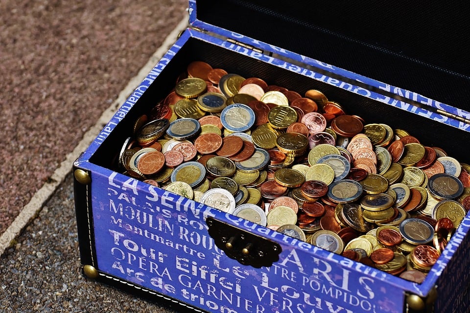 Baú azul decorado com nomes de pontos turísticos de Paris repleto de moedas de euro de diferentes valores e cores, representando economia, finanças e acumulação de dinheiro.