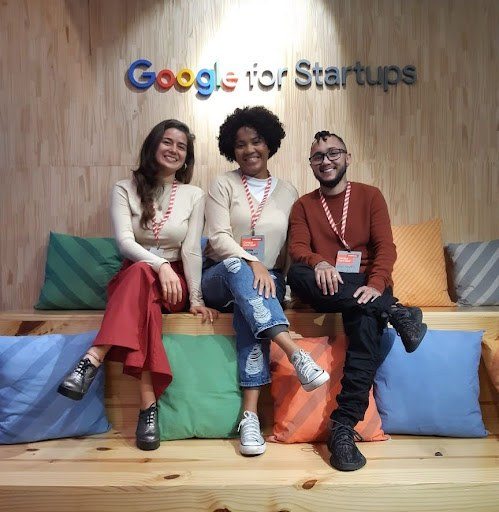 Foto com três pessoas, duas mulheres e um homem, sentados em um banco com almofadas. Na parede de fundo, escrito 