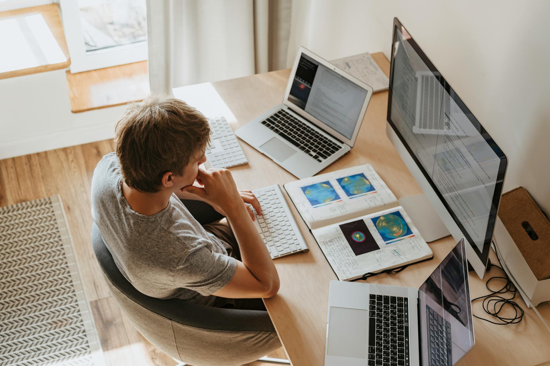 Visão aérea de um jovem sentado a uma mesa com um computador desktop, dois laptops abertos, um teclado separado e um caderno com anotações científicas, ilustrando um espaço de trabalho dedicado ao estudo de inteligência artificial na educação.