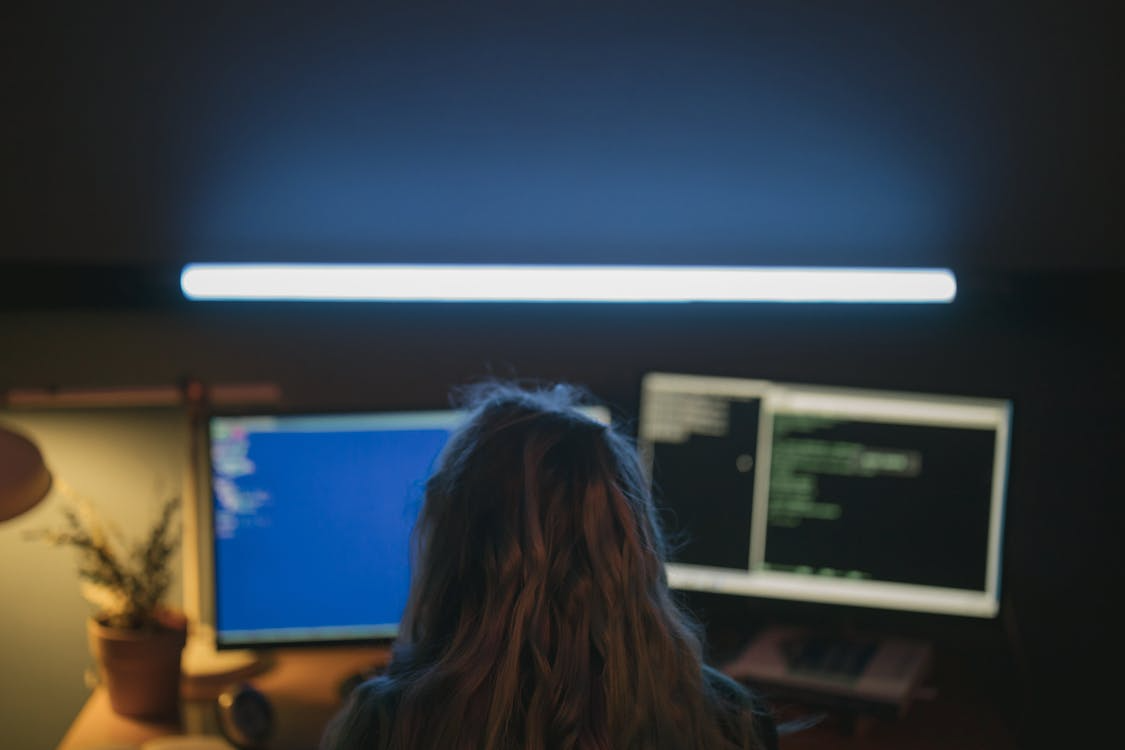 Pessoa engenheira de software vista de costas, trabalhando em um ambiente com pouca luz e múltiplos monitores, focada em programação e no desenvolvimento de sistemas.