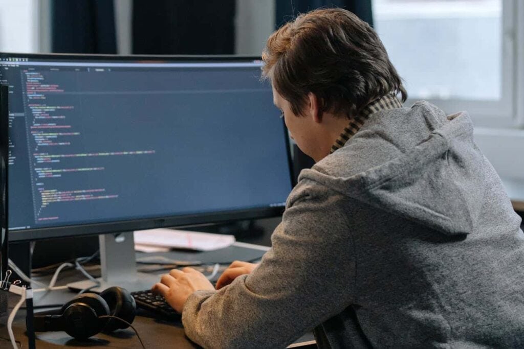 Desenvolvedor focado olhando para um monitor que exibe código-fonte em um editor, com fones de ouvido na mesa. Representa a programação assistida por IA e a produtividade em desenvolvimento.