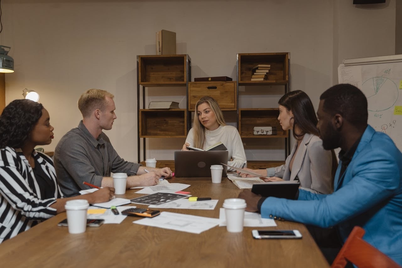 Cinco profissionais em volta de uma mesa de madeira em uma sala de reunião com iluminação quente. Uma mulher ao centro fala enquanto segura um caderno aberto. Os colegas ao redor ouvem atentamente, com copos de café, papéis e um laptop sobre a mesa.