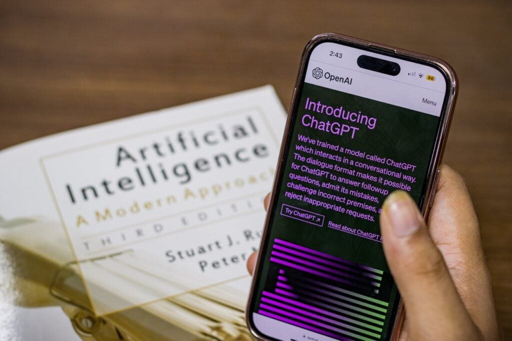 Uma mão segura um smartphone exibindo a página "Introducing ChatGPT" da OpenAI. Ao fundo, levemente desfocado, está o livro clássico de Stuart Russell e Peter Norvig, "Artificial Intelligence: A Modern Approach".