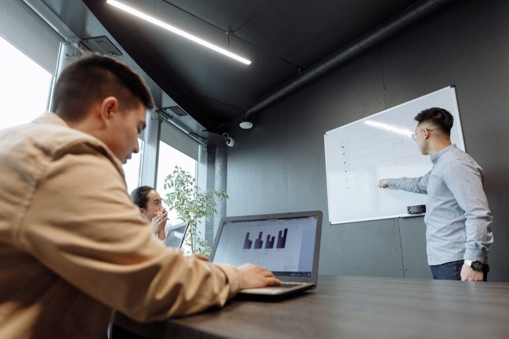 Ambiente de escritório moderno com três pessoas. Um homem em pé aponta para um quadro branco com gráficos desenhados, enquanto outro colega em primeiro plano observa e utiliza um notebook. Uma mulher ao fundo acompanha a explicação.