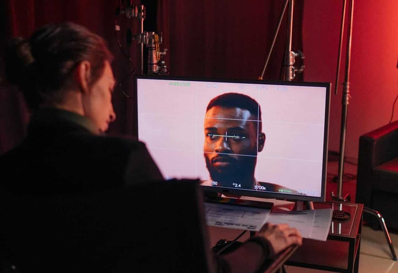 Uma pessoa sentada à frente de um notebook em um ambiente escurecido. Na tela, um software de edição de vídeo exibe diversas marcações de vídeo (timeline) e uma prévia de um vídeo de um homem em destaque.