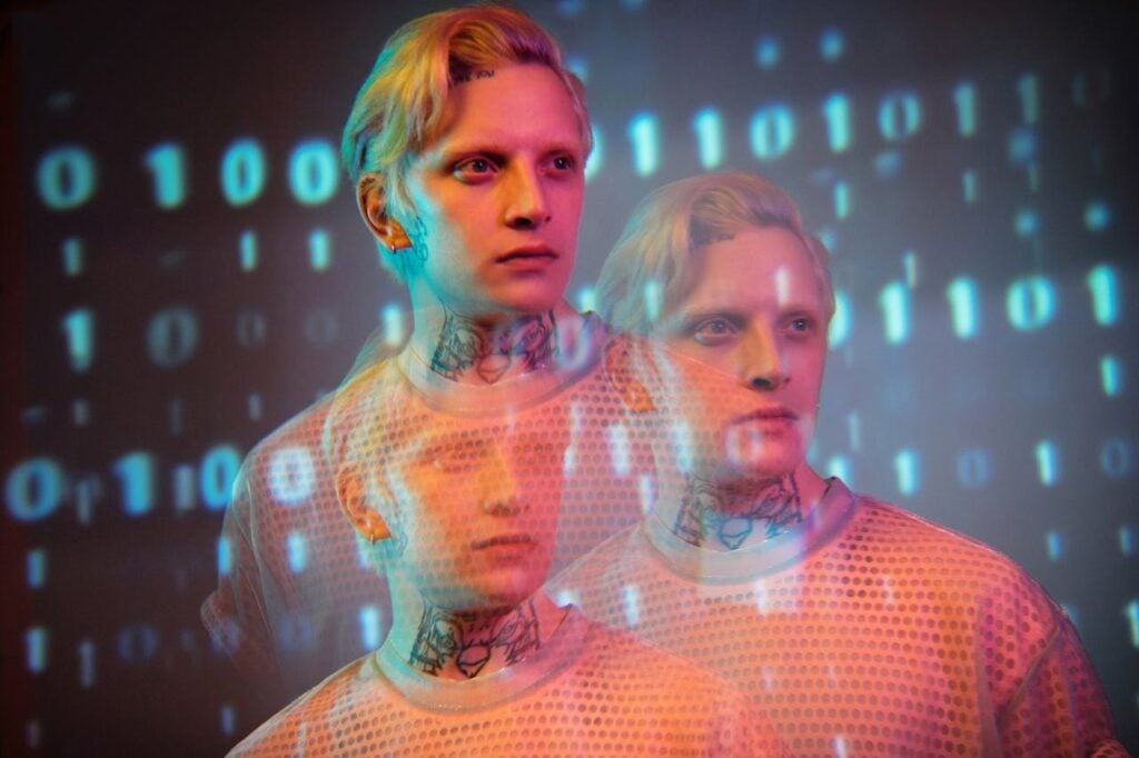 Três figuras humanas estilizadas com efeito digital translúcido, sobrepostas a código binário em azul e laranja, simbolizando personas geradas por inteligência artificial