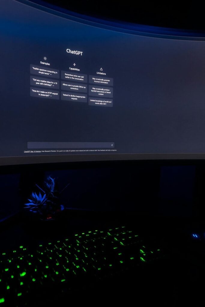Close-up de um monitor curvo exibindo a tela inicial do ChatGPT no escuro. As informações de capacidades e limitações do modelo estão centralizadas na tela. Abaixo, um teclado mecânico com iluminação em LED verde brilha intensamente, criando um contraste com o ambiente escuro.