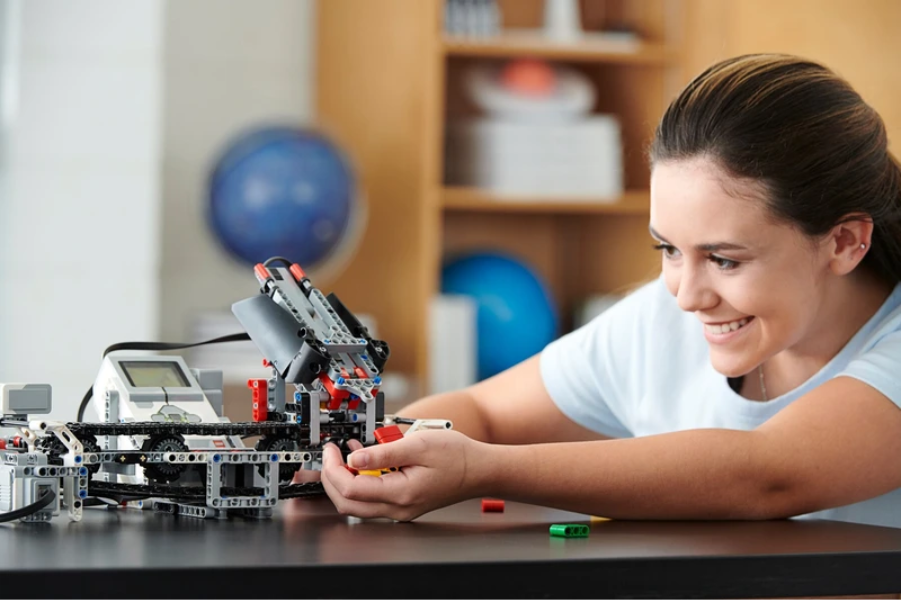 Pessoa sorrindo enquanto monta e ajusta um robô feito com peças de montar (LEGO Technic) sobre uma mesa. O robô possui engrenagens, sensores e uma pequena tela conectada. Ao fundo, há prateleiras com objetos desfocados, incluindo uma bola azul. 