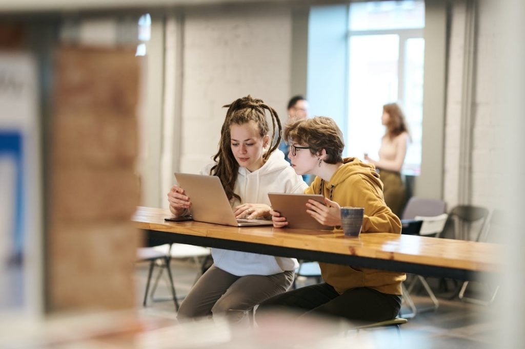 Duas pessoas jovens sentadas lado a lado em uma mesa de madeira clara em um ambiente de coworking. Uma delas utiliza um laptop prateado e a outra um tablet, ambas concentradas nas telas, representando uma equipe analisando métricas de visibilidade digital.