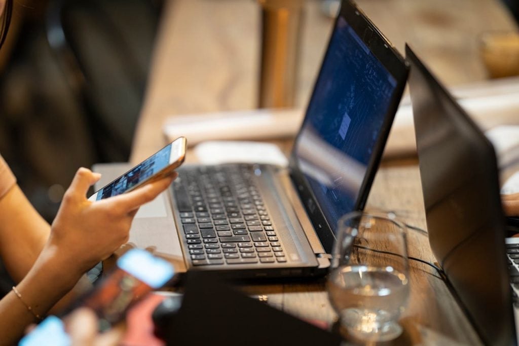 Close-up das mãos de uma pessoa segurando um smartphone enquanto trabalha em um laptop aberto sobre uma mesa de madeira. A composição sugere o uso de múltiplos pontos de acesso para interação com mecanismos de busca e IAs. 