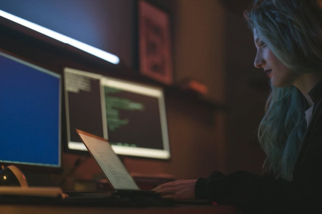 Perfil de uma mulher jovem com cabelos claros ondulados, usando óculos de grau, concentrada diante de três telas em um ambiente escurecido. A tela do notebook em primeiro plano e os dois monitores ao fundo exibem códigos de programação e interfaces de sistemas em tons de verde e azul.