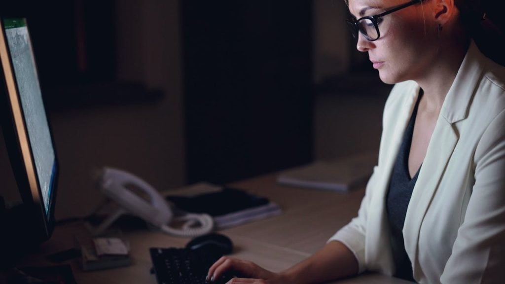 Mulher de óculos e blazer branco sentada à mesa de trabalho durante a noite. Ela olha atentamente para um monitor que ilumina seu rosto. Sobre a mesa, veem-se um teclado preto, um mouse e um telefone fixo branco ao fundo. A iluminação é focada, sugerindo um ambiente de escritório ou home office noturno.