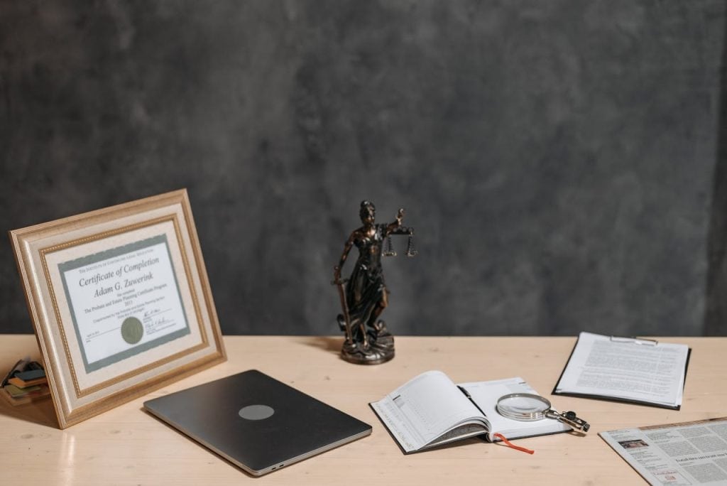 Mesa de trabalho de madeira clara com um laptop fechado, um certificado de conclusão emoldurado à esquerda e uma pequena estátua de bronze da Dama da Justiça ao centro. Sobre a mesa, há também um livro aberto com uma lupa em cima e papéis de petição organizados, representando um ambiente jurídico moderno e preparado para a pesquisa.