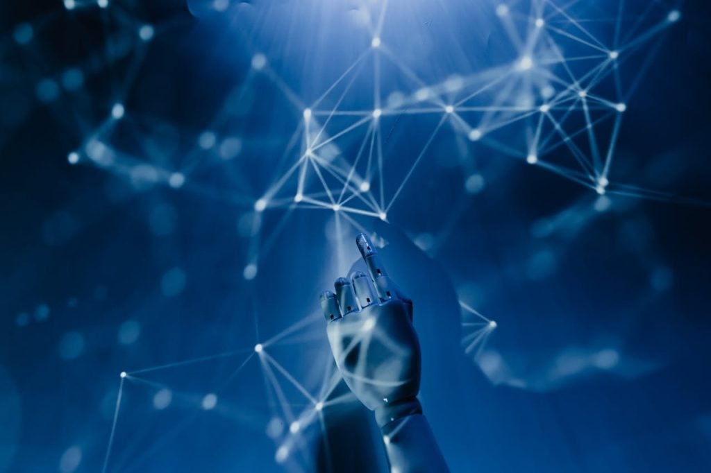 Imagem conceitual de tecnologia com fundo azul escuro, mostrando uma mão robótica branca apontando para cima em direção a uma rede de pontos conectados por linhas brilhantes, representando redes neurais e inteligência artificial.