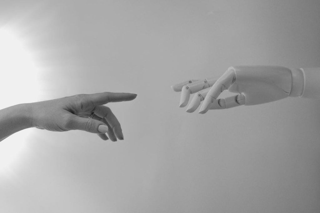 Fotografia em preto e branco mostrando uma mão humana à esquerda estendendo o dedo indicador em direção a uma mão robótica à direita, que também estende o dedo, em uma referência visual à união entre humanos e tecnologia.