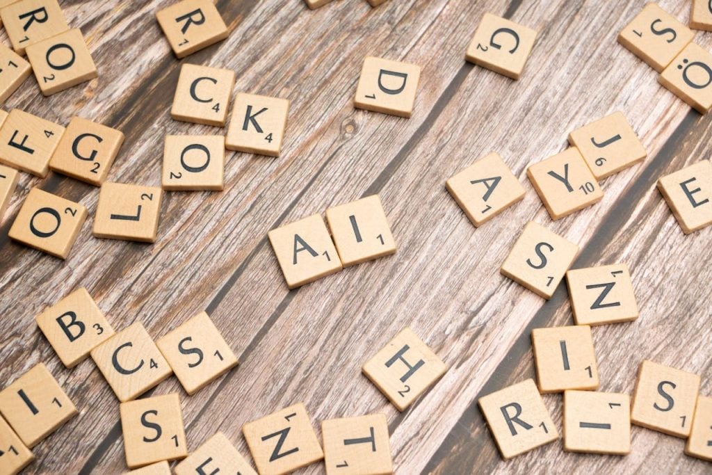 Diversas peças de madeira de um jogo de palavras (estilo Scrabble) espalhadas sobre uma superfície de madeira clara. No centro, as letras formam em destaque a sigla "AI" (Inteligência Artificial).