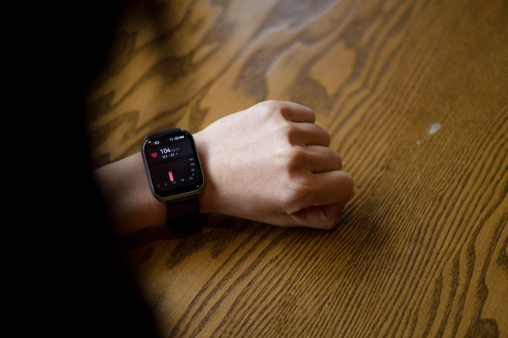  Visão em ângulo superior focando no pulso de uma pessoa com a mão espalmada sobre uma mesa de madeira rústica com veios marcados. No pulso, ela usa um relógio inteligente (smartwatch) preto com tela digital ligada marcando o horário de "11:02 AM", exibindo um ícone de coração vermelho e a medição de batimentos cardíacos atual de "104 bpm", além de um gráfico de barras na parte inferior da interface.