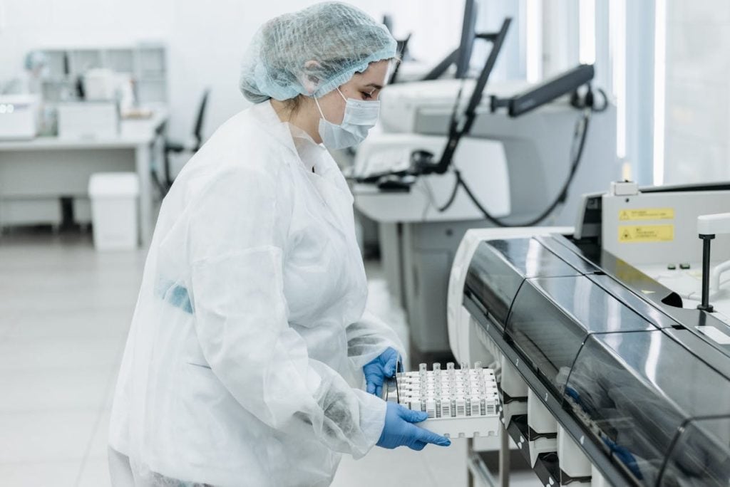 Mulher usando jaleco ou avental de proteção branco por cima da roupa, touca cirúrgica azul-clara e máscara facial protetora branca. Ela está de perfil em um laboratório médico automatizado com maquinários hospitalares ao fundo, segurando firmemente com as duas mãos cobertas por luvas cirúrgicas azuis um suporte de plástico branco carregado com múltiplos tubos de ensaio de coleta de sangue transparentes e organizados.