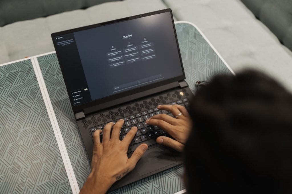 Visão em ângulo superior por cima do ombro de uma pessoa com cabelos escuros, focando em suas mãos digitando no teclado de um notebook preto sobre uma mesa com estampa geométrica cinza e branca. A tela do notebook está ligada e exibe a interface escura do ChatGPT, mostrando as três colunas padrão de informações organizadas.