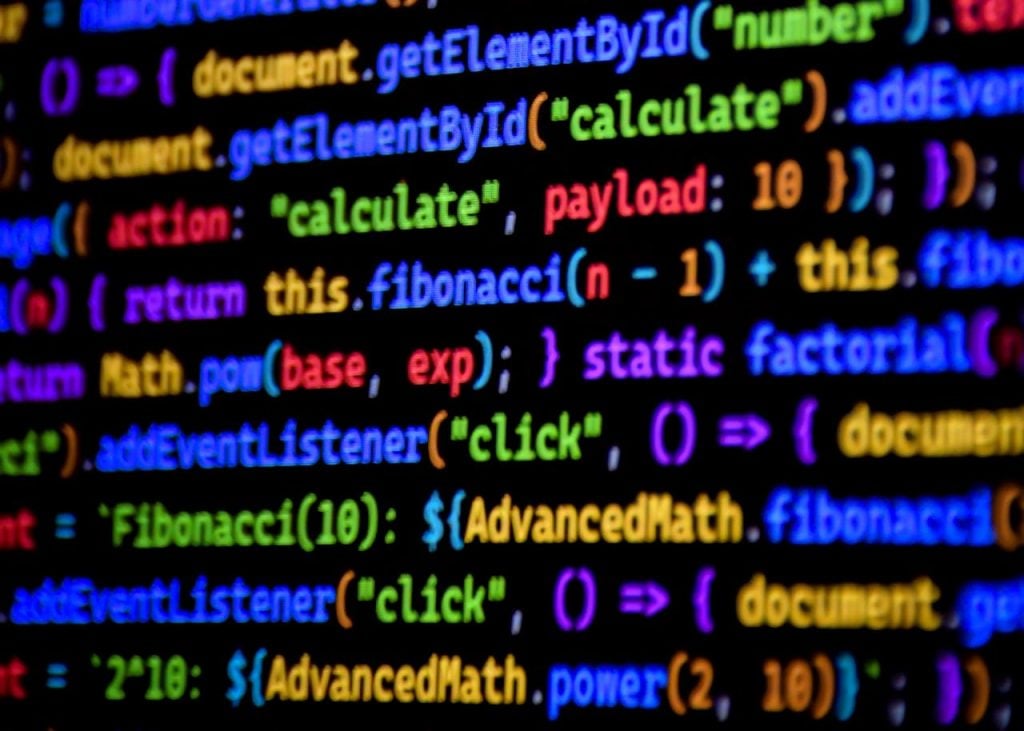 Close-up focado com fundo preto exibindo múltiplas linhas de código de programação JavaScript na tela de um computador. O texto do código está altamente colorido com sintaxe destacada em tons brilhantes de azul, verde, amarelo, roxo e laranja, exibindo termos técnicos legíveis.