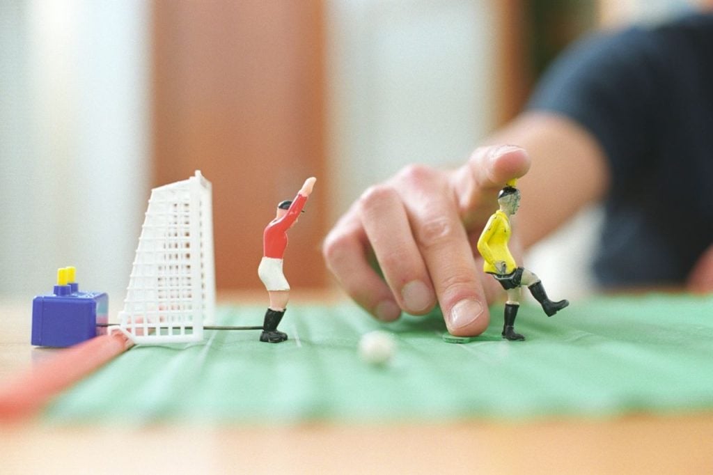 Visão aproximada e em ângulo baixo de pequenas réplicas de plástico de jogadores de futebol sobre um tabuleiro verde texturizado que simula um campo. Um boneco veste camisa vermelha com calção branco e outro veste camisa amarela com calção preto. Em segundo plano, uma mão humana estende o dedo indicador para tocar nas peças.