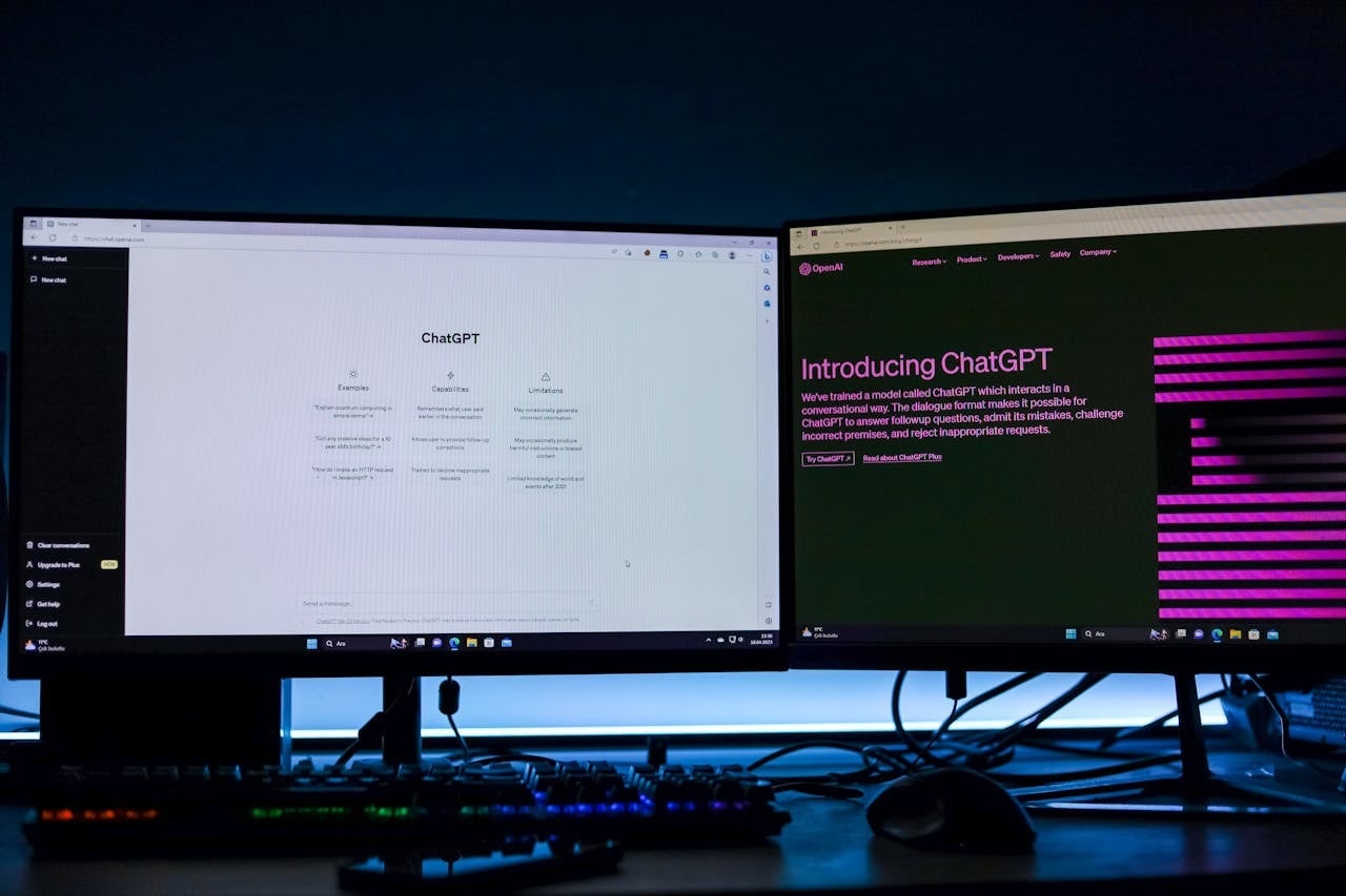 Duas telas de monitor lado a lado em uma mesa escura com teclado iluminado. A tela da esquerda exibe a interface inicial em modo claro do ChatGPT com suas colunas de exemplos e capacidades. A tela da direita mostra o site da OpenAI com a página de introdução ao ChatGPT em fundo escuro.