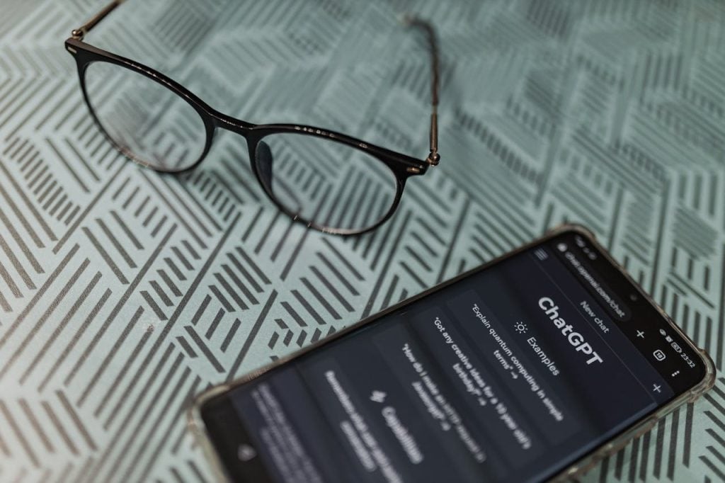 Visão aproximada de um smartphone deitado sobre uma mesa com estampa geométrica verde e branca. A tela do celular exibe o aplicativo do ChatGPT em modo escuro. Um par de óculos de grau com armação preta está pousado logo acima do aparelho.