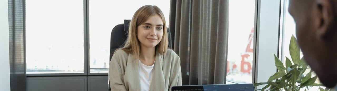 Uma jovem mulher está sentada em um escritório moderno, participando de uma entrevista de emprego. Em primeiro plano, vê-se apenas a silhueta das costas e da cabeça de um recrutador negro, que analisa um documento em uma prancheta de papel. Entre os dois, há um laptop aberto sobre a mesa.