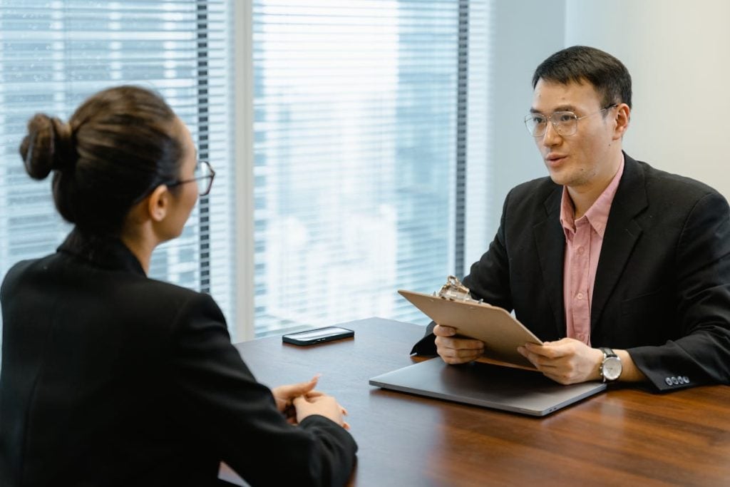 Visão em ângulo médio de uma entrevista corporativa. À direita, um homem de óculos e blazer preto conversa com uma candidata de cabelos presos em um coque, vista de costas à esquerda. O recrutador segura uma prancheta de madeira e um laptop fechado repousa sobre a mesa de madeira polida.