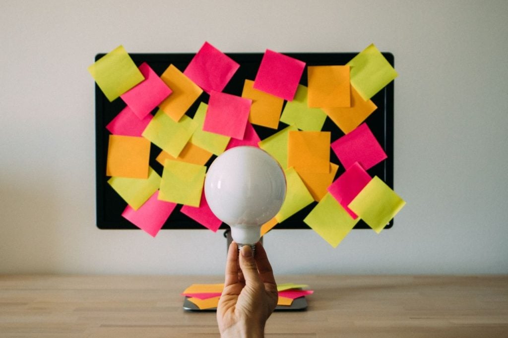 Uma mão segura uma lâmpada branca acesa em primeiro plano, centralizada sobre uma mesa de madeira clara. Ao fundo, um monitor de computador está completamente coberto por dezenas de blocos de notas adesivas (post-its) coloridos em tons vibrantes de rosa, amarelo e laranja.