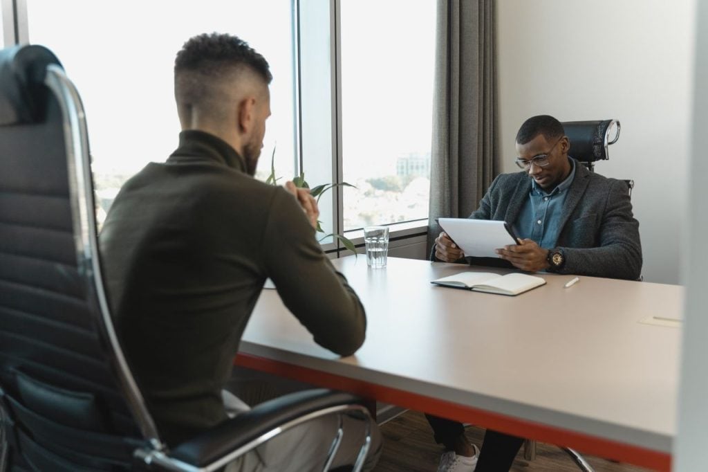 Em uma sala de reuniões corporativa, um recrutador usa óculos e blazer cinza enquanto analisa atentamente uma folha de currículo presa a uma prancheta. À esquerda, um candidato de blusa verde-escura está sentado de costas, aguardando em uma cadeira de escritório preta.
