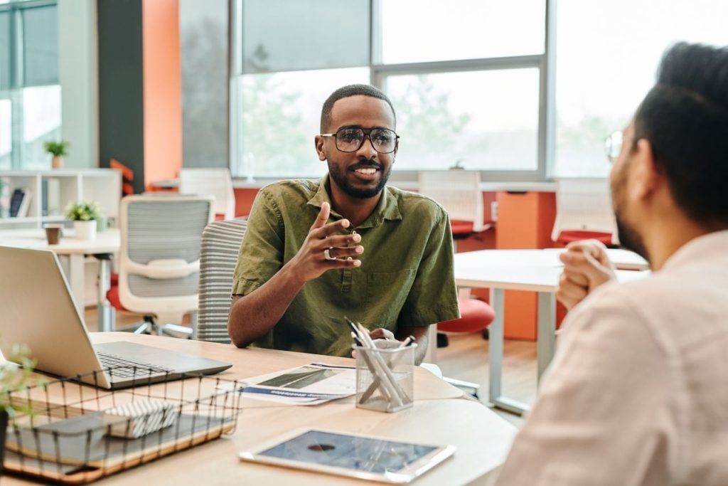 Em um escritório iluminado, um jovem homem está sentado à mesa conversando e gesticulando com a mão esquerda. Ele está de frente para um profissional de camisa branca visto de costas em primeiro plano. Sobre a mesa, há um notebook aberto e alguns papéis de relatório.