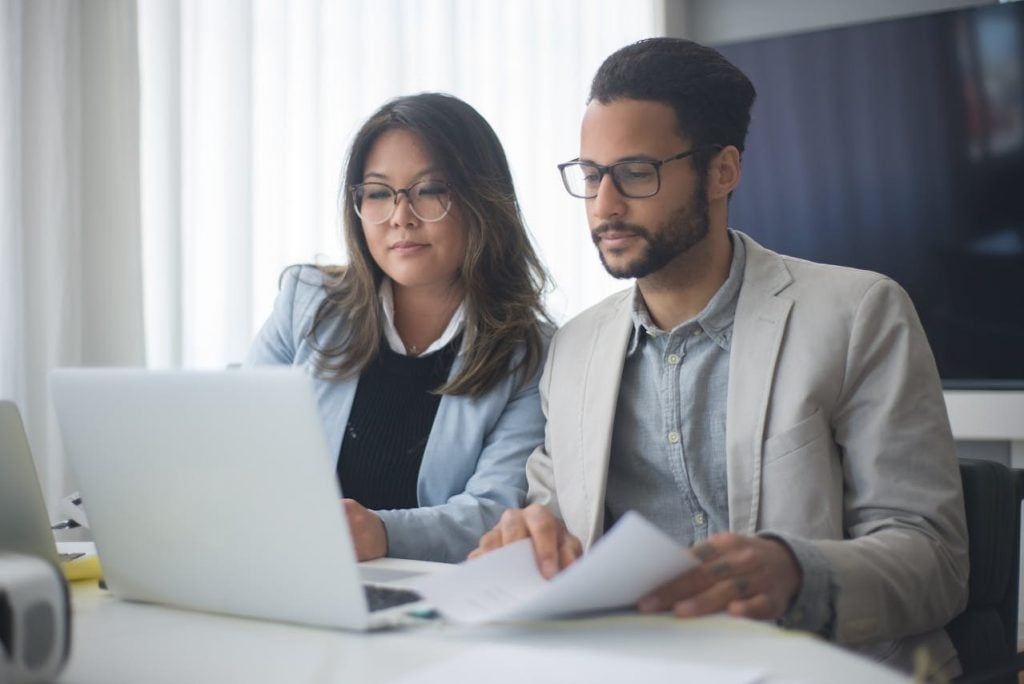 Uma mulher de óculos e blazer azul claro e um homem de barba, óculos e blazer bege trabalham lado a lado focados na tela de um notebook branco. O homem segura algumas folhas de papel impresso enquanto analisa as informações digitalizadas ao lado de sua colega de equipe.