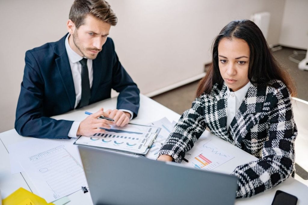 Homem de terno escuro e gravata observa atentamente uma mulher ao seu lado trabalhando em um notebook. Sobre a mesa branca, há relatórios impressos com gráficos de pizza e de barras. A mulher usa um blazer xadrez em preto e branco e foca o olhar na tela do computador