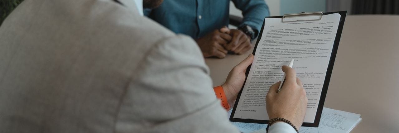 Close das mãos de um homem com blazer bege segurando uma prancheta que exibe um contrato comercial ou proposta de trabalho. Ele usa uma caneta stylus para apontar ou assinar um campo do documento. No fundo desfocado, um colega usando camisa jeans o observa atentamente durante a reunião.