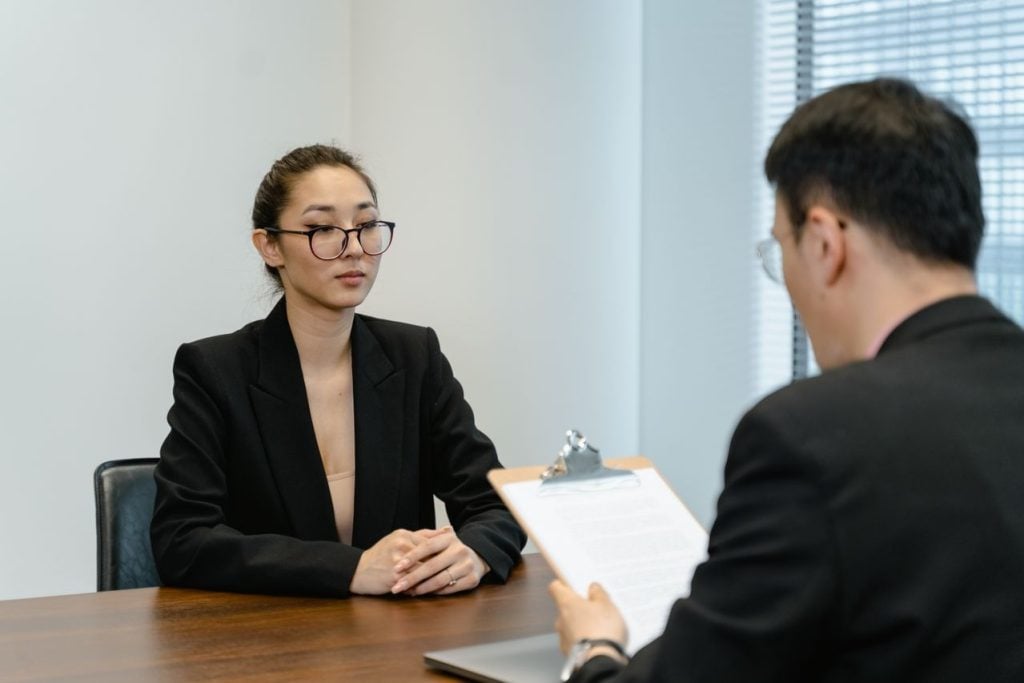 Uma mulher de óculos e blazer e um homem, óculos e blazer bege fazem uma entrevista lado a lado focados na tela de um notebook branco. O homem segura algumas folhas de papel impresso enquanto analisa as informações digitalizadas.