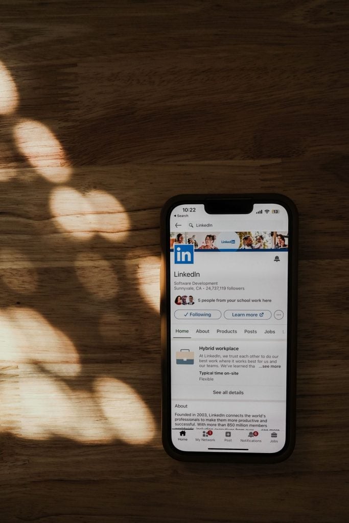 Smartphone sobre uma mesa de madeira exibindo a página do LinkedIn, com luz natural projetando sombras suaves ao redor do aparelho.